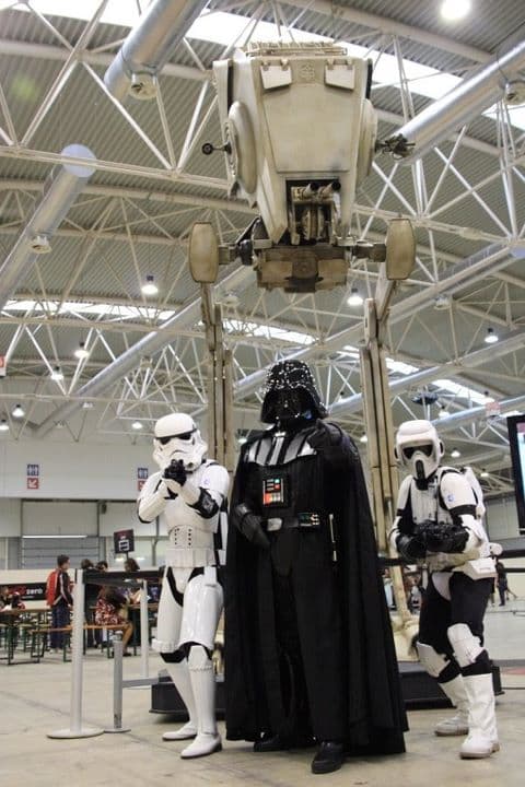: Replica in scala 1:1 dell'AT-ST in esposizione, con cosplayer di Star Wars in primo piano che ne mostrano l'imponente altezza.