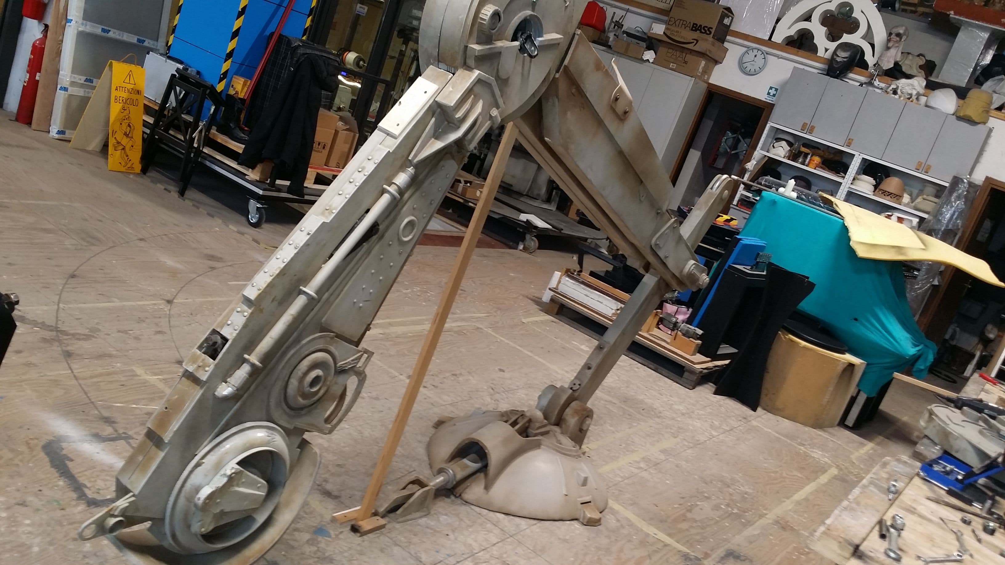 Struttura articolata di una gamba meccanica dell'AT-ST posata a terra e sostenuta da un'asta di supporto nel laboratorio scenografico.